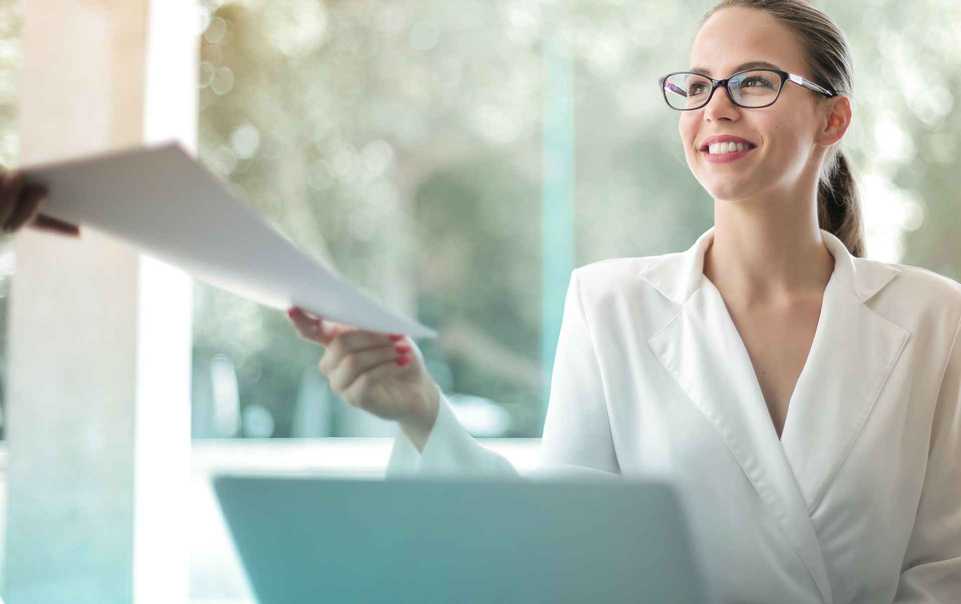 Low angle of successful female executive manager in classy style sitting at table with laptop in contemporary workplace and passing documents to colleague